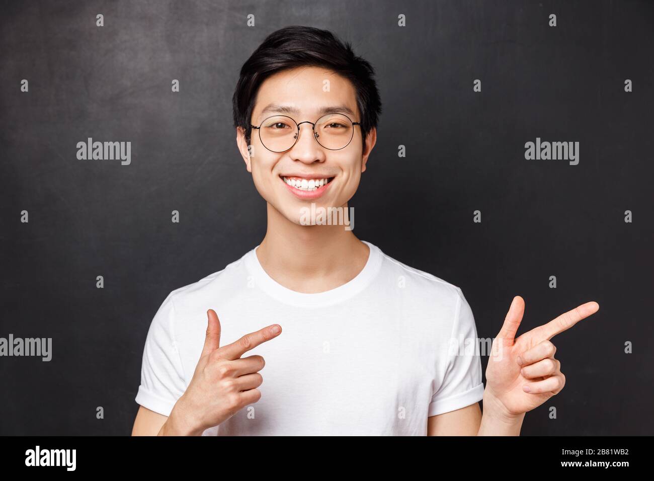 Close-up portrait of friendly smiling asian man in white t-shirt and glasses looking at camera confident, pointing fingers right at promo or company Stock Photo