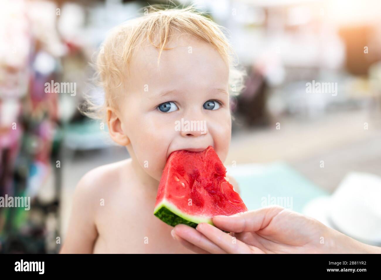 Mother feeding cute adorable caucasian blond little toddler boy slice of fresh sweet tasty watermelon at vacation on sea or ocean beach. Child biting Stock Photo