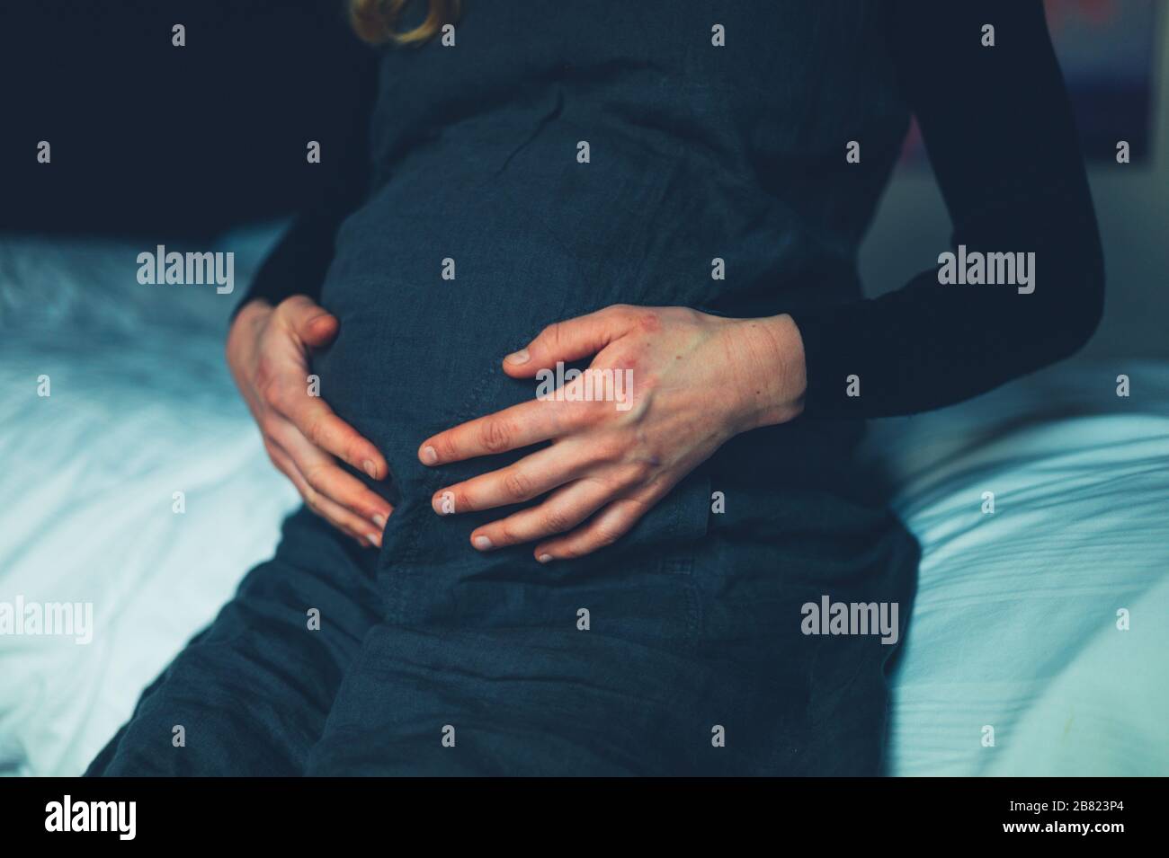 A pregnant woman with eczema on her hands is sitting on a bed feeling her stomach Stock Photo