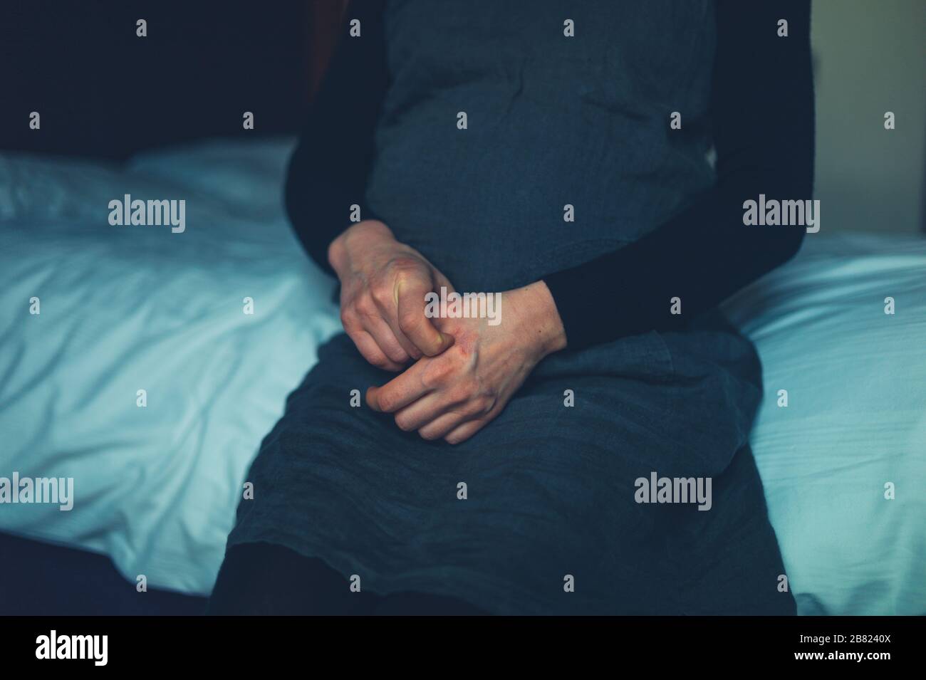 A pregnant woman with eczema is sitting on a bed scratching her hand Stock Photo