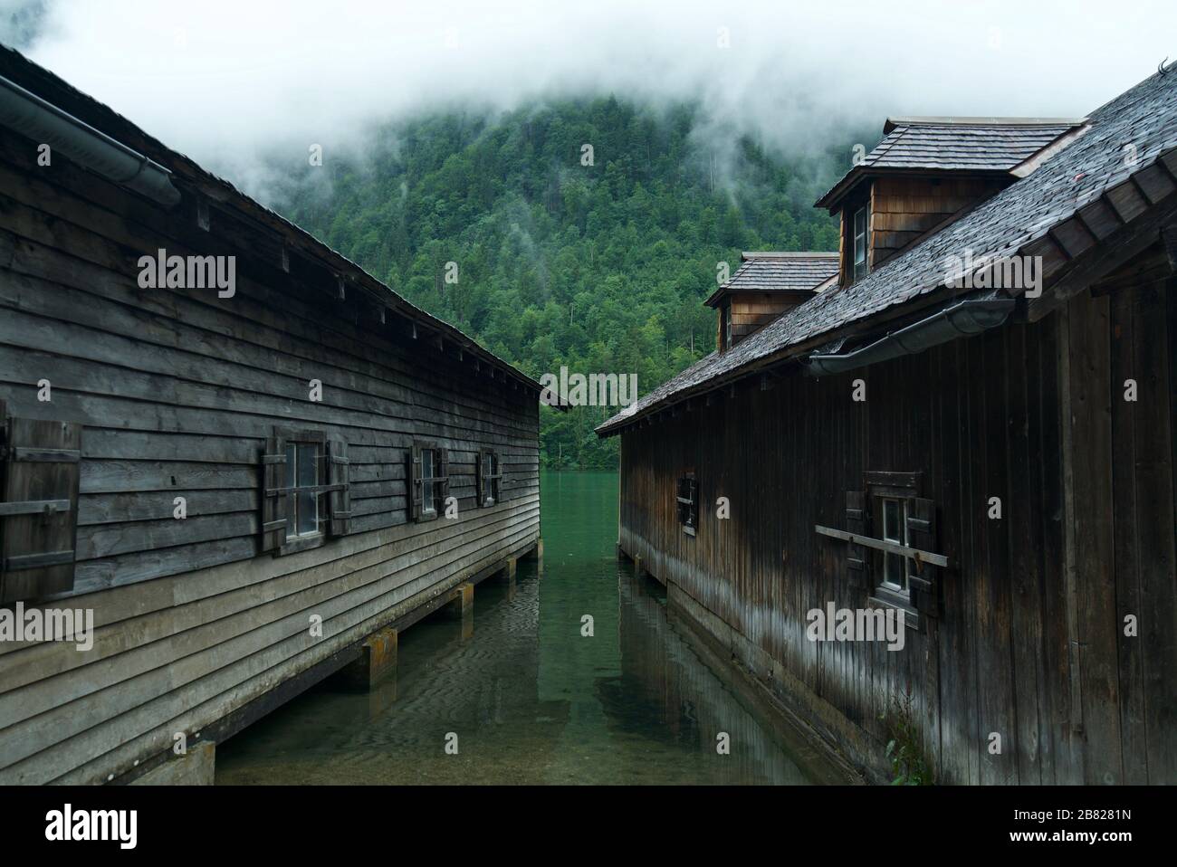 Boathouse / Boathouses by the water surrounded by mountains and clouds at Lake Königssee (Berchtesgaden National Park). Wooden cabins by the lake. Stock Photo