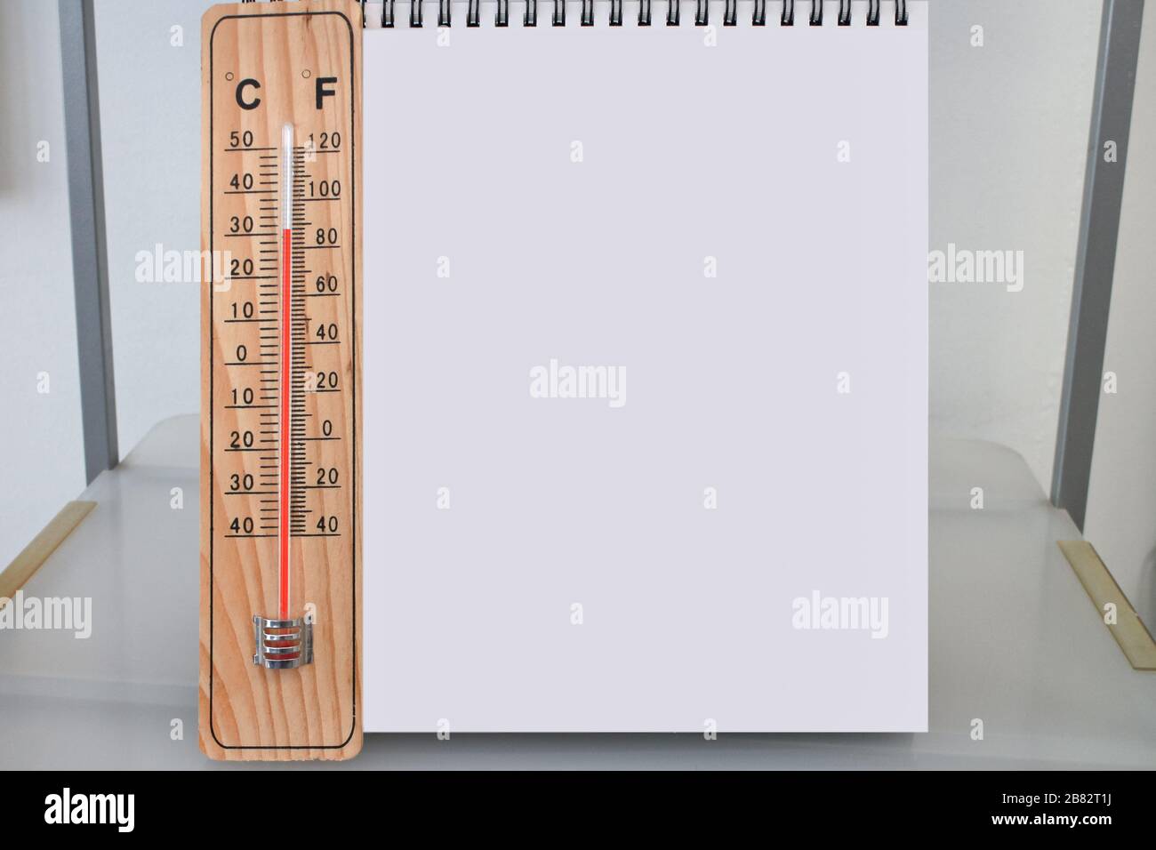 Celsius and Fahrenheit indoor wooden thermometer with an empty desk calendar or blank notepad on a shelf on a hot summer day, mockup and template Stock Photo
