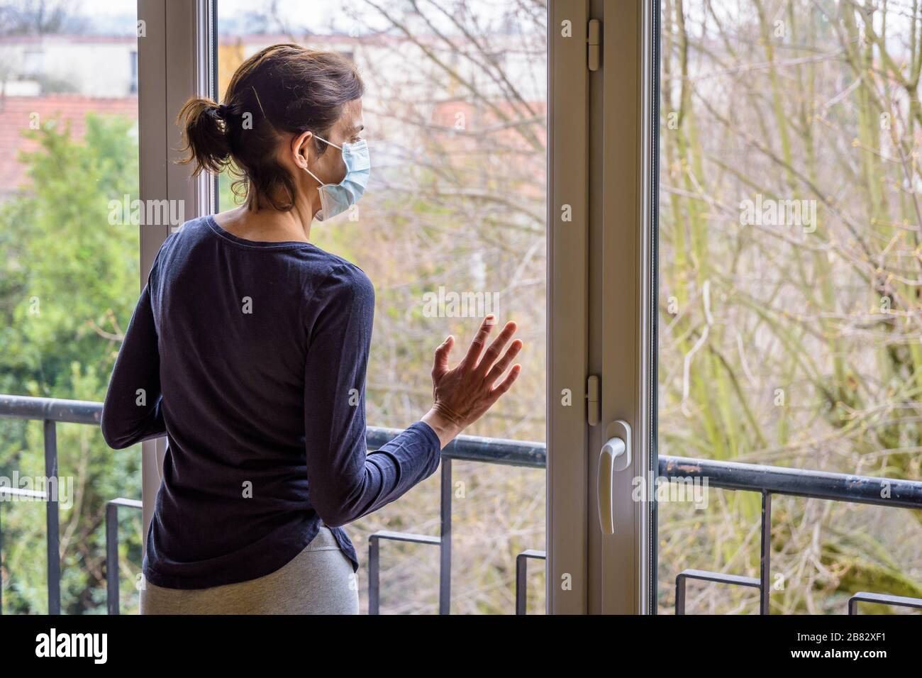 A woman under quarantine at home, wearing a medical face mask and a casual outfit, is standing idle in front of a closed window, staring into space. Stock Photo