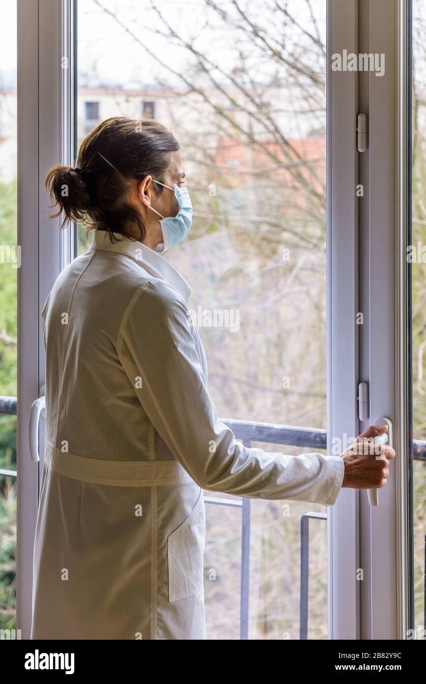 A nurse wearing a medical face mask and a white coat, is standing idle in front of a closed window, staring into space. Stock Photo