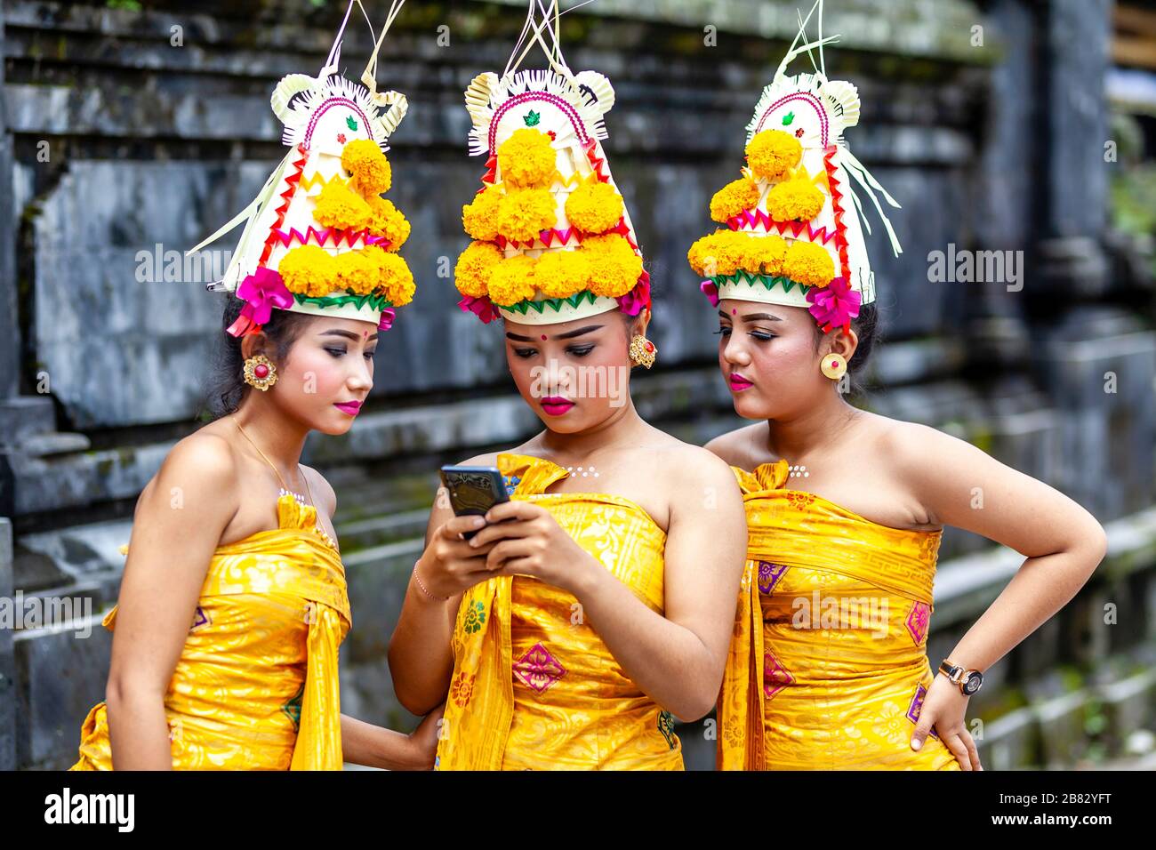 Young Balinese Hindu Females Looking At A Mobile Phone (Cellphone) At The Batara Turun Kabeh Ceremony, Besakih Temple, Bali, Indonesia. Stock Photo