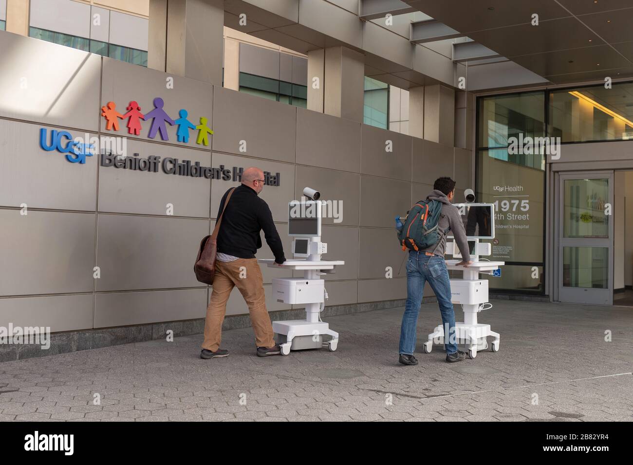 Two staff members wheel Amwell telemedicine carts into the entrance of the University of California San Francisco (UCSF) Benioff Children's Hospital in Mission Bay, San Francisco, California during an outbreak of the COVID-19 coronavirus, March 16, 2020. As a result of the outbreak, patients are increasingly being asked to conduct telemedicine appointments to avoid infecting healthcare workers. () Stock Photo