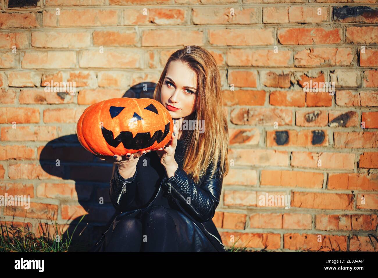 modern Halloween woman. Attractive witch holding pumpkin 'trick or treat'. Woman dressing in leather jacket and black skirt. October halloween. bricks Stock Photo