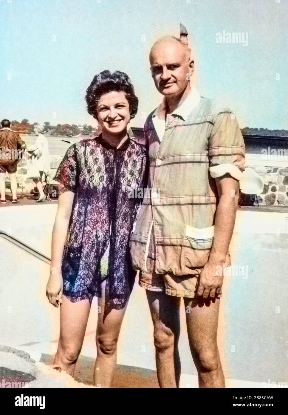 Mature American tourist couple posing in front of a swimming pool, with woman wearing a bathing suit and cover-up and man wearing shorts and a shirt, 1970. Note: Image has been digitally colorized using a modern process. Colors may not be period-accurate. () Stock Photo
