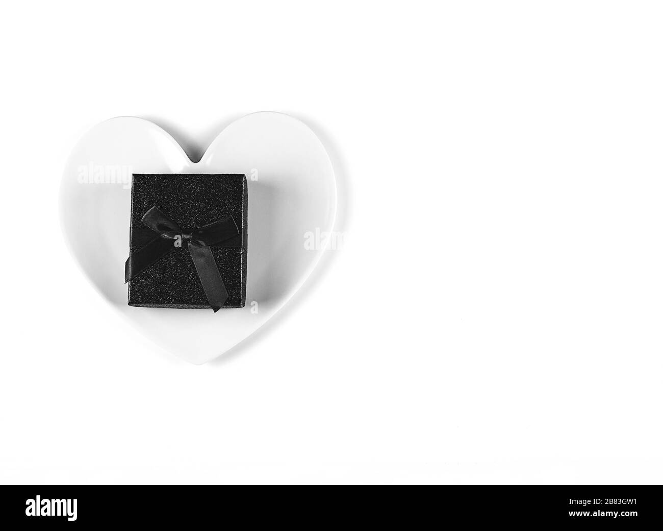 Black Gift box with a bow, a white plate in the shape of a heart on white background. Minimalist Black concept Stock Photo
