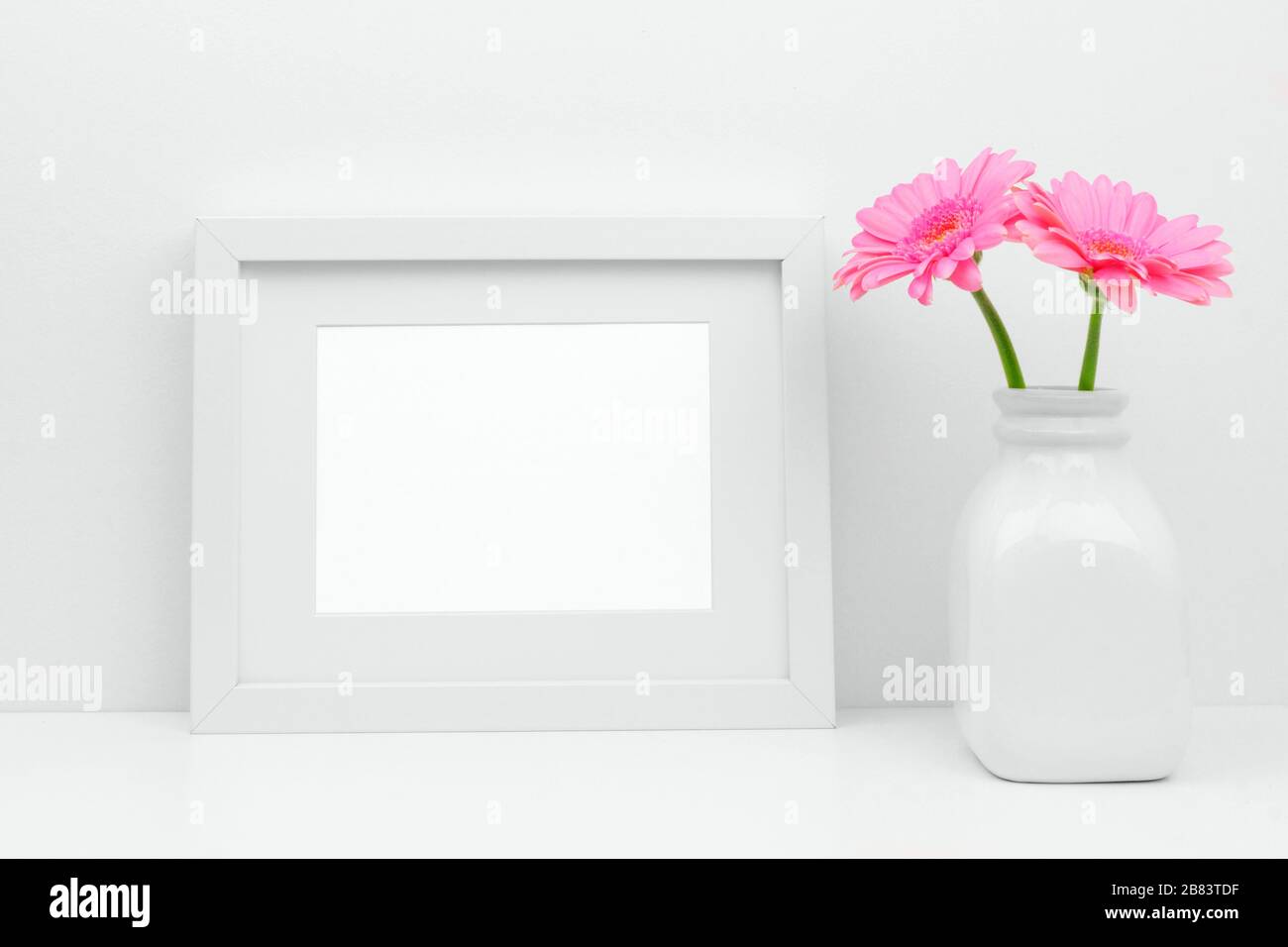 Mock up white frame and pink daisy flowers in vase on a shelf or desk. White color scheme. Landscape frame orientation. Stock Photo