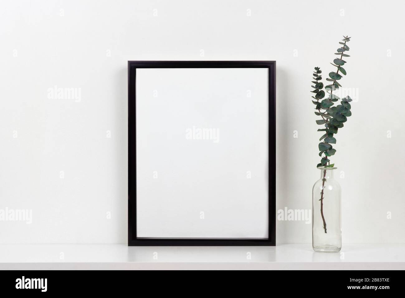 Mock up black frame with vase of branches on a shelf or desk. White shelf and wall. Portrait frame orientation. Stock Photo