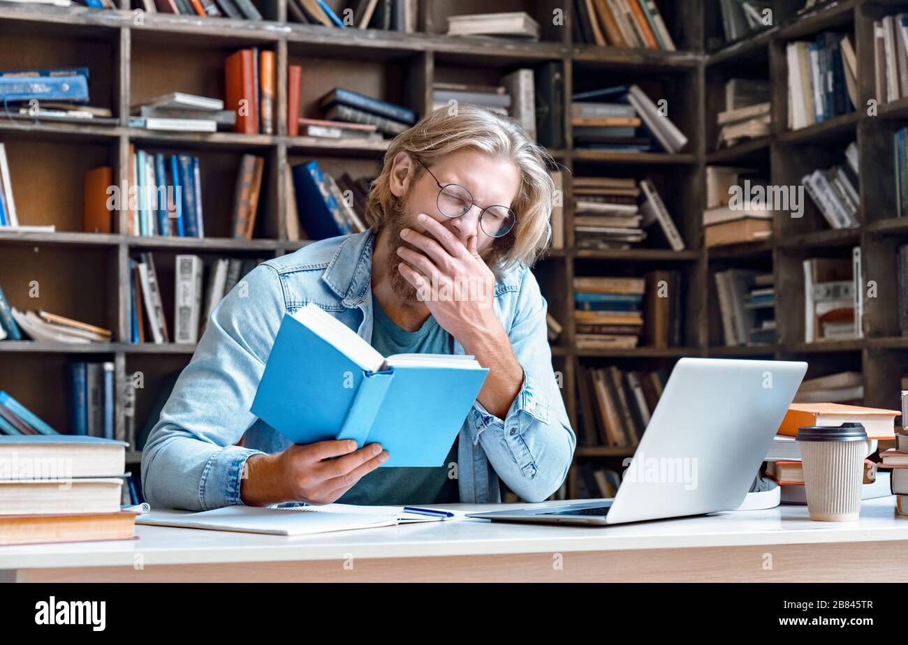 Bored tired guy student yawning reading book studying in library. Stock Photo