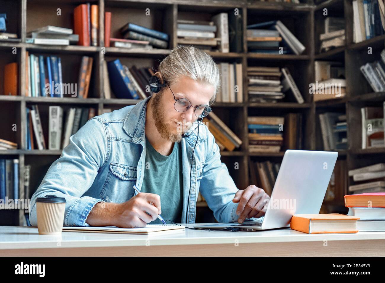 Focused man student wear headset study online make notes in library. Stock Photo