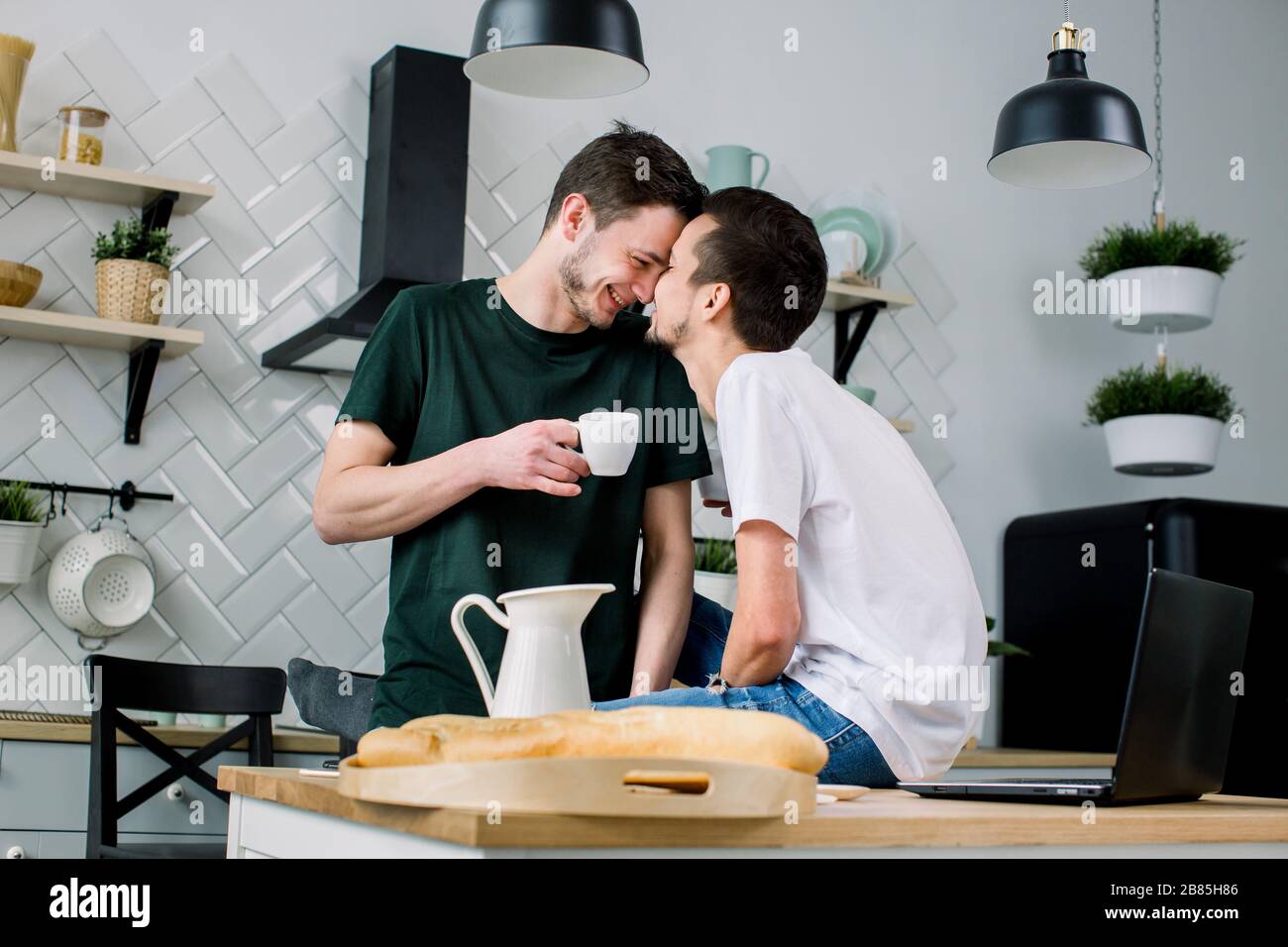 Happy gay couple having breakfast in kitchen and using laptop. Amorous gay men kissing each other, face to face while spending leisure at home. Stock Photo