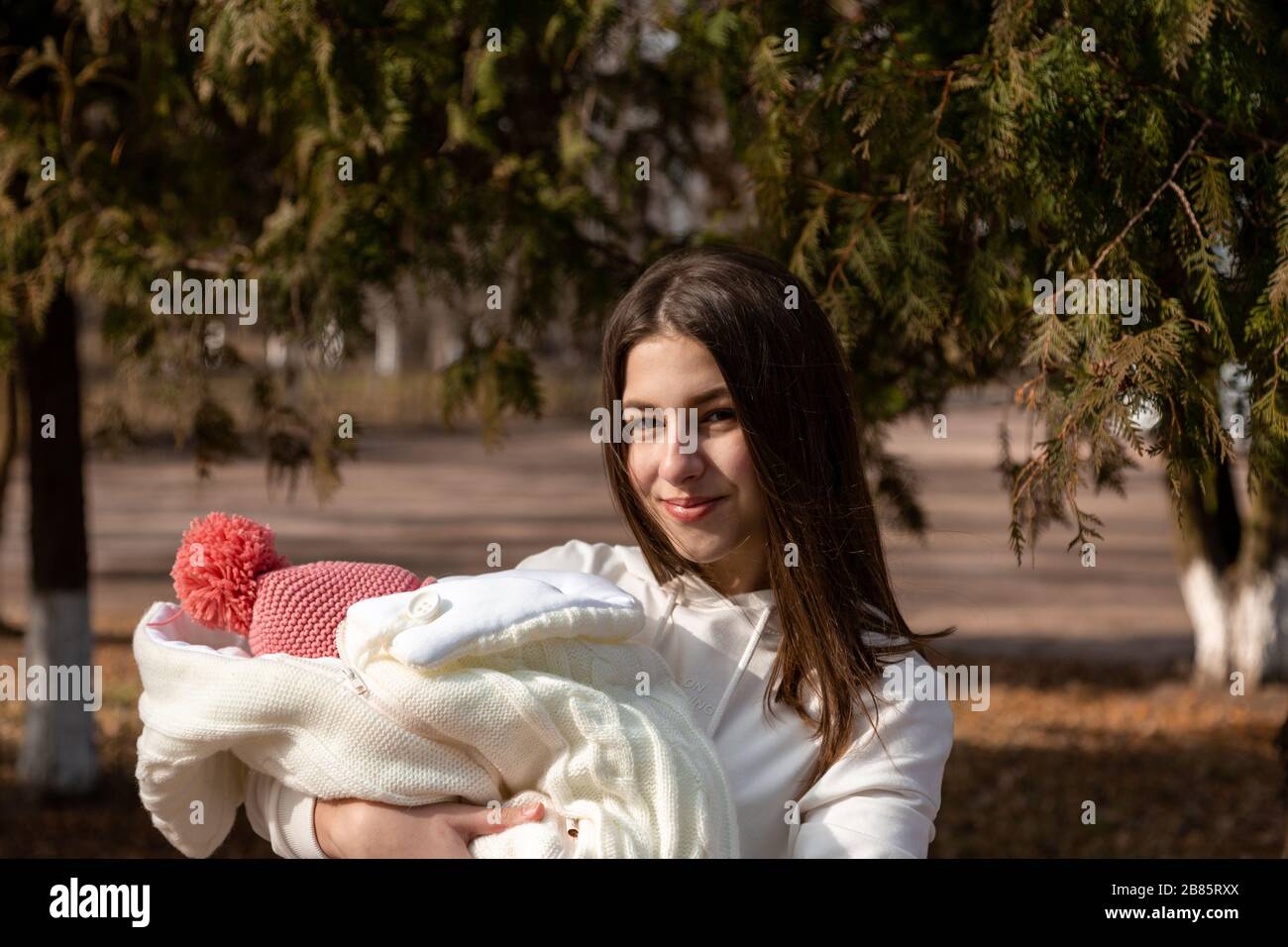 Pretty young woman holding a newborn baby in her arms. They are standing outside on a sunny day around the Christmas tree. Stock Photo