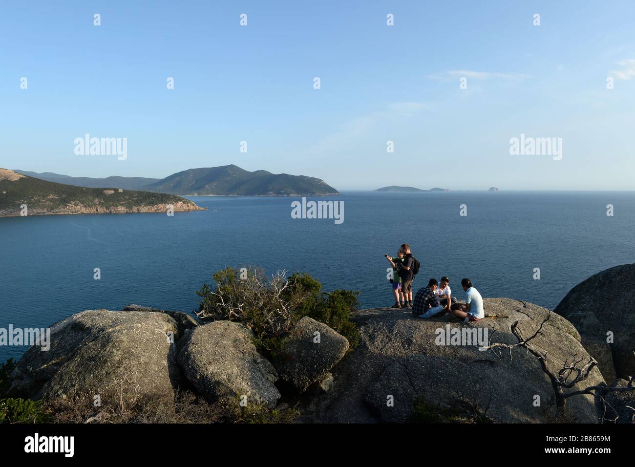 Group of young men  play cards on hilltop overlooking bay while a young couple take photos of the view Stock Photo