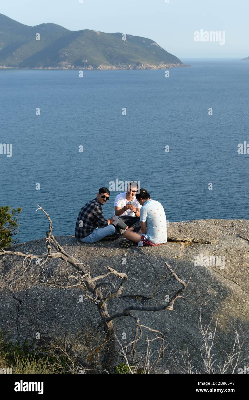 Three friends play a game of cards on top of a granite boulder overlooking the sea at Pillar Point, Australia Stock Photo
