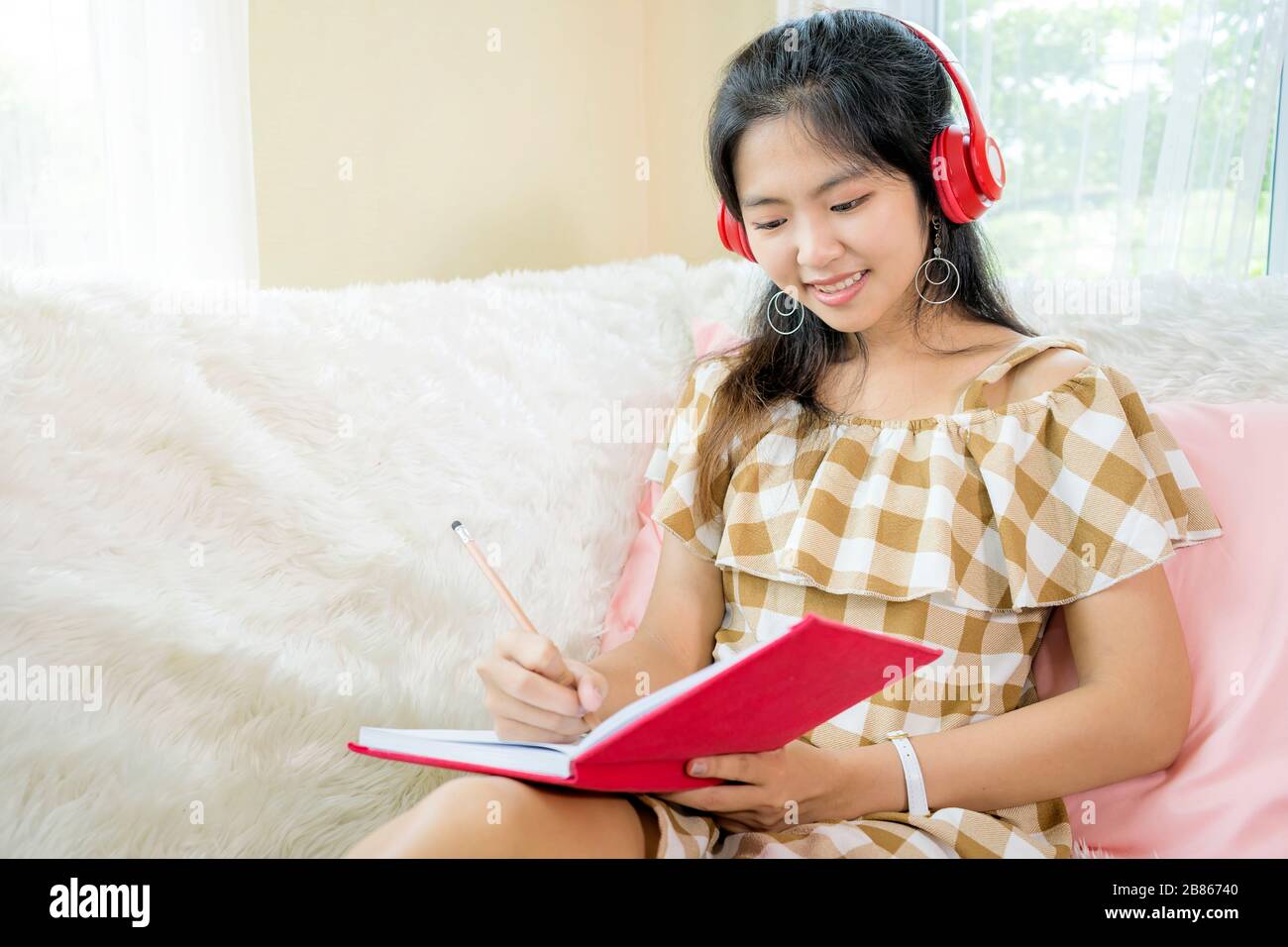 Asian women, pretty face was writing a note to that, she was wearing red headphones to listen to music, relax the mind, sitting on a white sofa in her Stock Photo