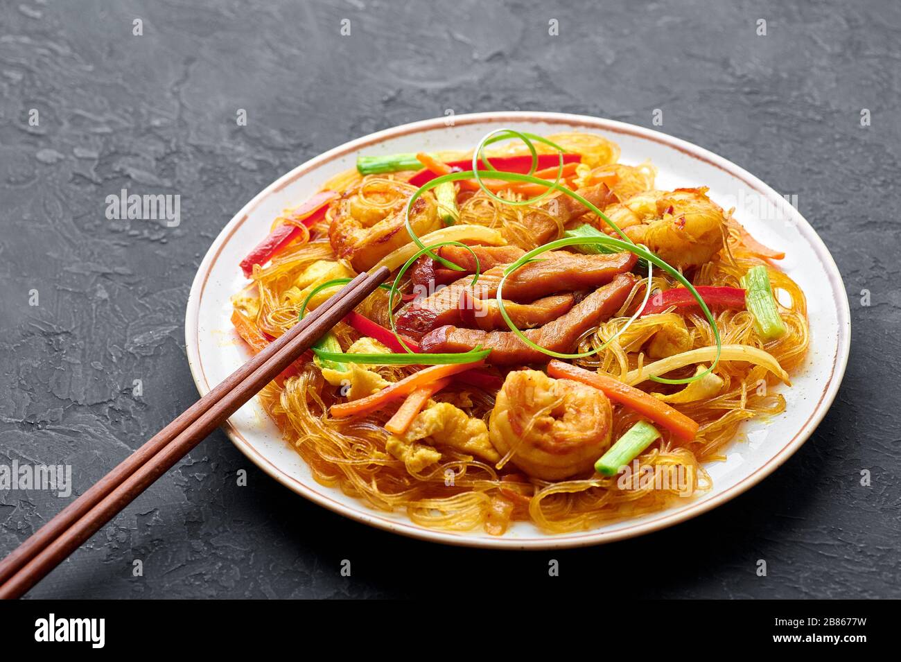 Singapore Mei Fun in white plate on dark slate background. Singapore Noodles is chinese cuisine dish with rice noodles, prawns, char siu pork, carrot, Stock Photo