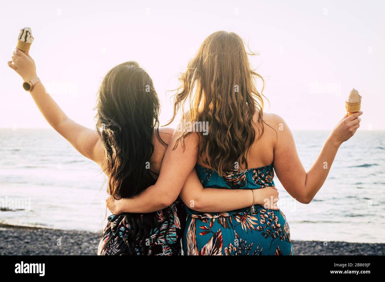 Rear back view of couple of women friends hug and enjoy together the summer eating an ice cream and looking at the blue sea in background - concept of Stock Photo