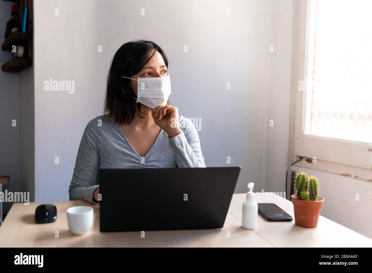 young woman wearing protective face mask and with hand sanitizer in home office looking worried and stressed out of the window due to corona virus Stock Photo