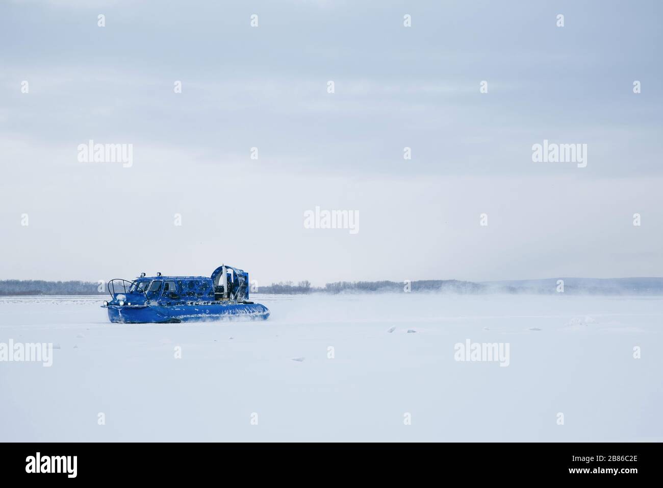 A blue hovercraft rushes along the frozen ice of the Volga River. Behind her rises from the snow. Stock Photo