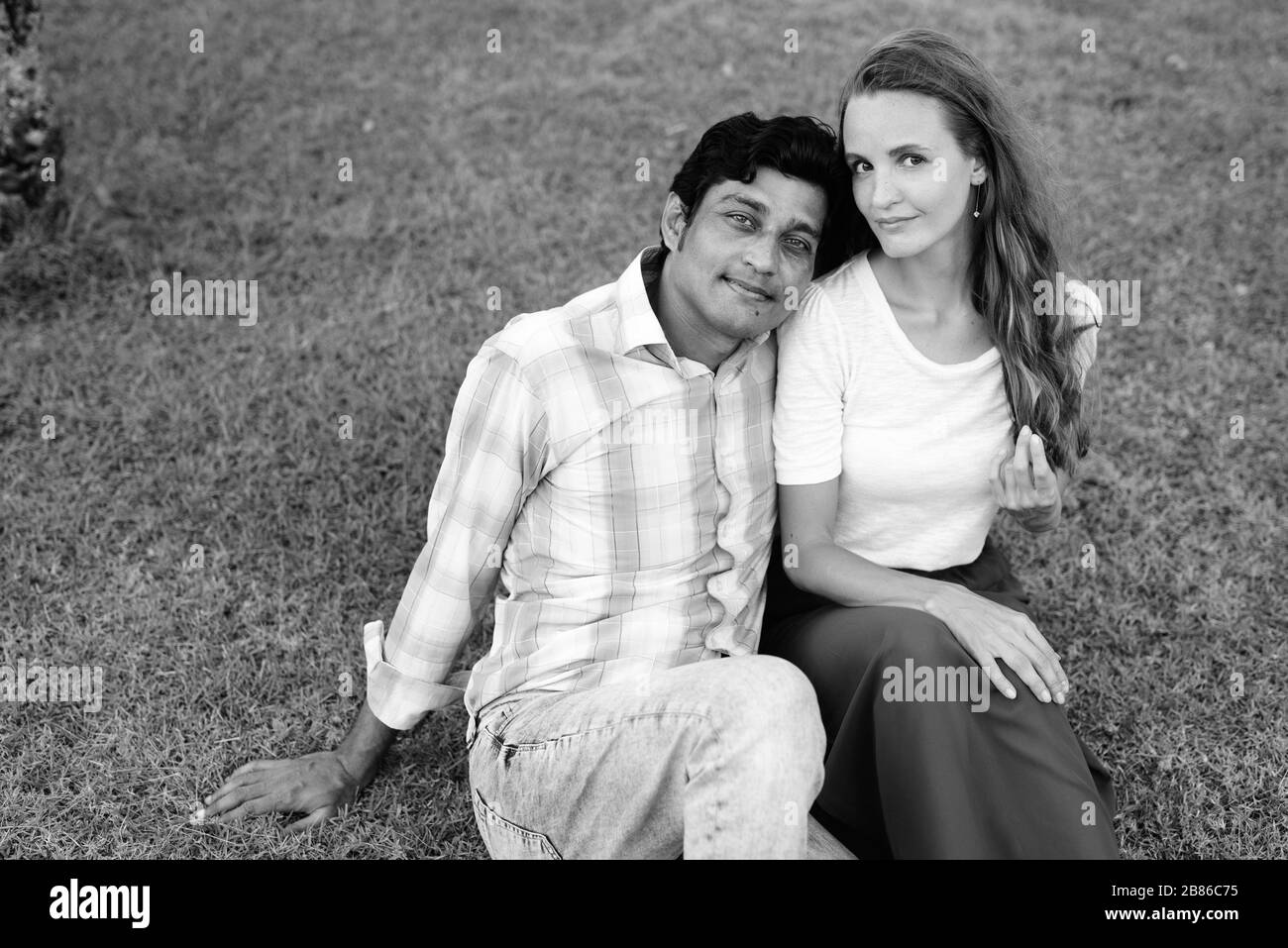 Multi ethnic couple sitting on the meadow in love with man leaning on the shoulder of woman at the park Stock Photo