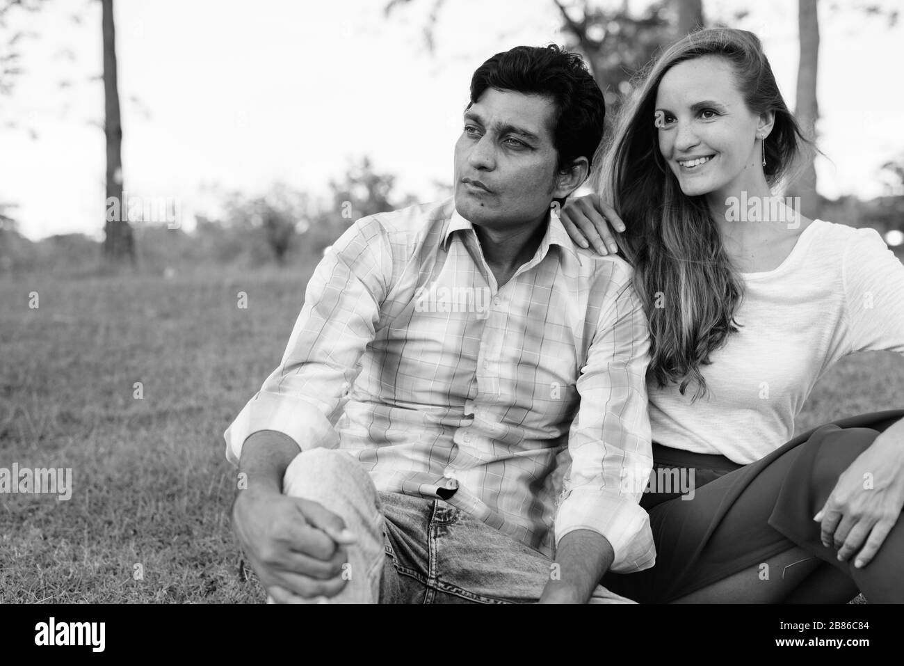 Happy multi ethnic couple smiling while thinking and sitting on the meadow in love at the park Stock Photo