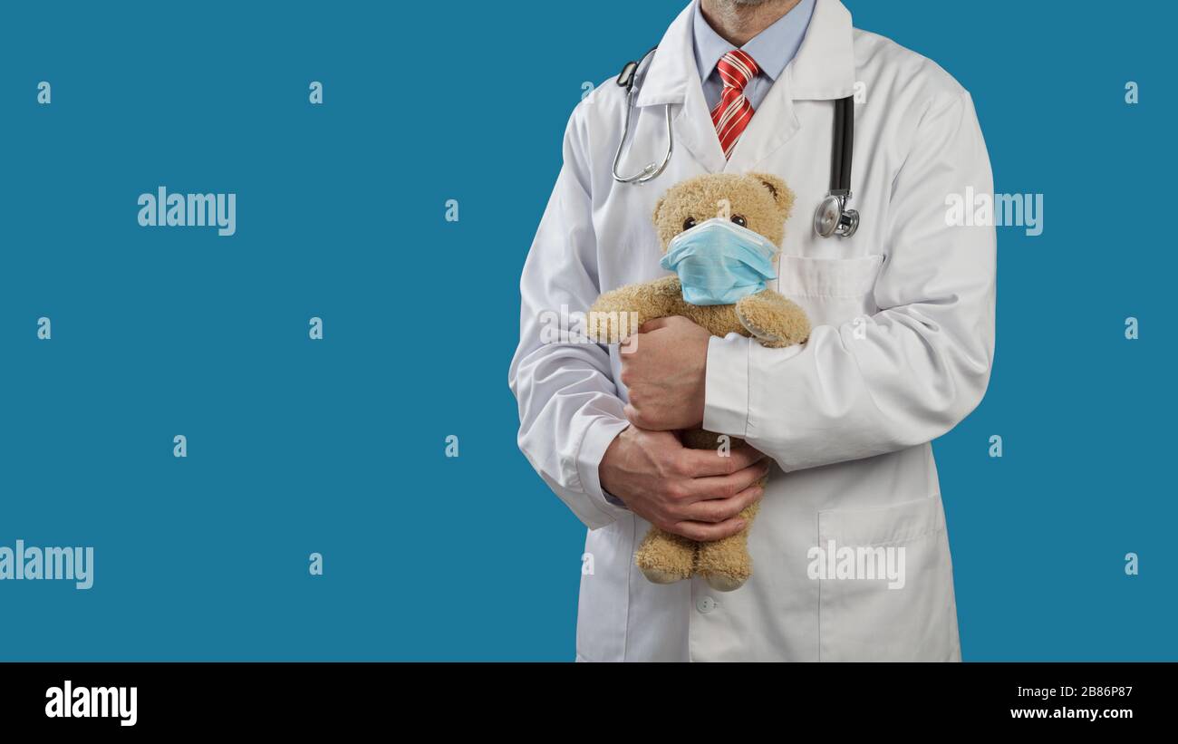 Medical doctor pediatrician with a stethoscope on shoulders holds a teddy bear in a medical mask. Stock Photo