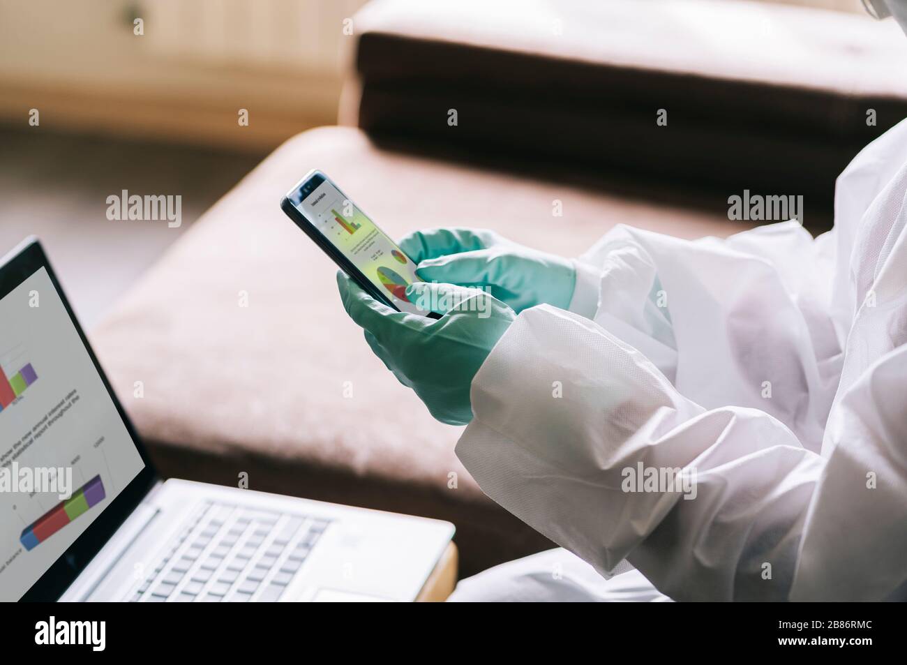 The hands of a person using the mobile phone with gloves and a safety suit against a pandemic or virus Stock Photo