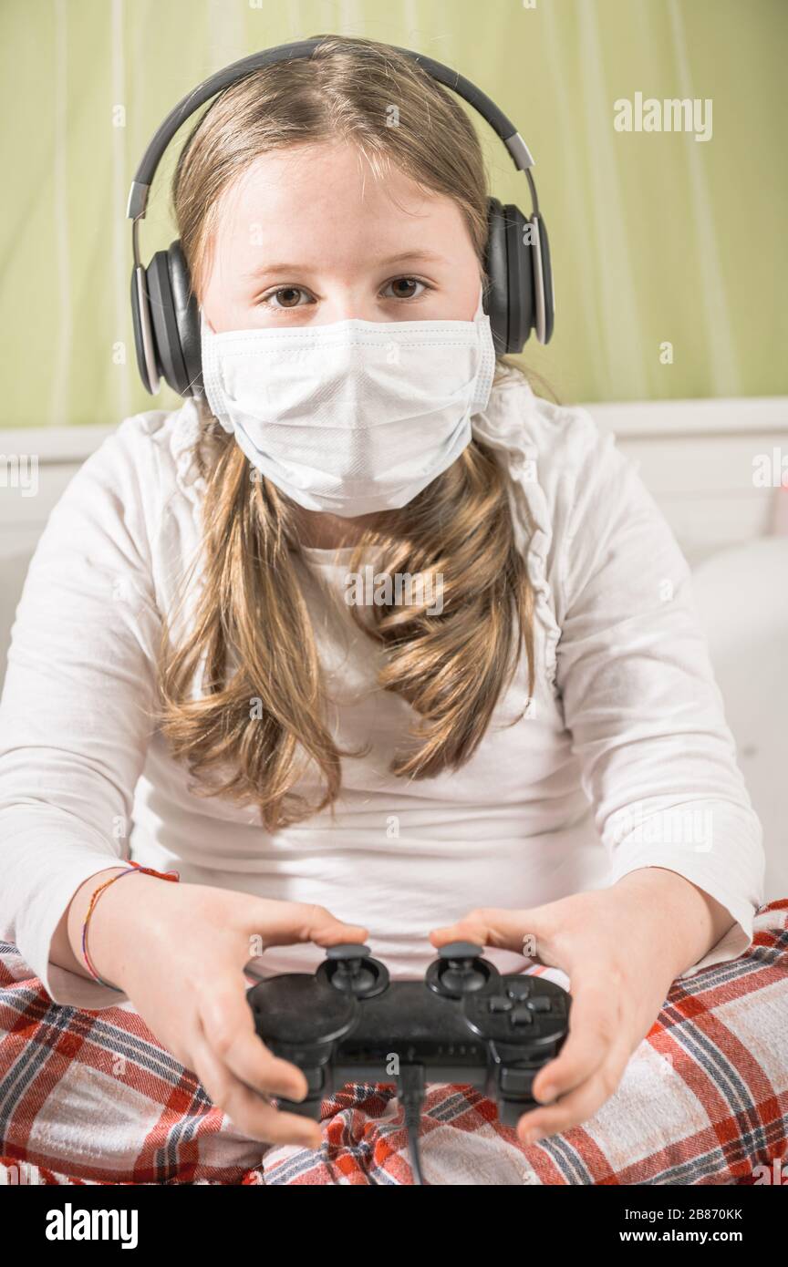 Girl in a mask playing on a game console during quarantine on the bed. Stock Photo