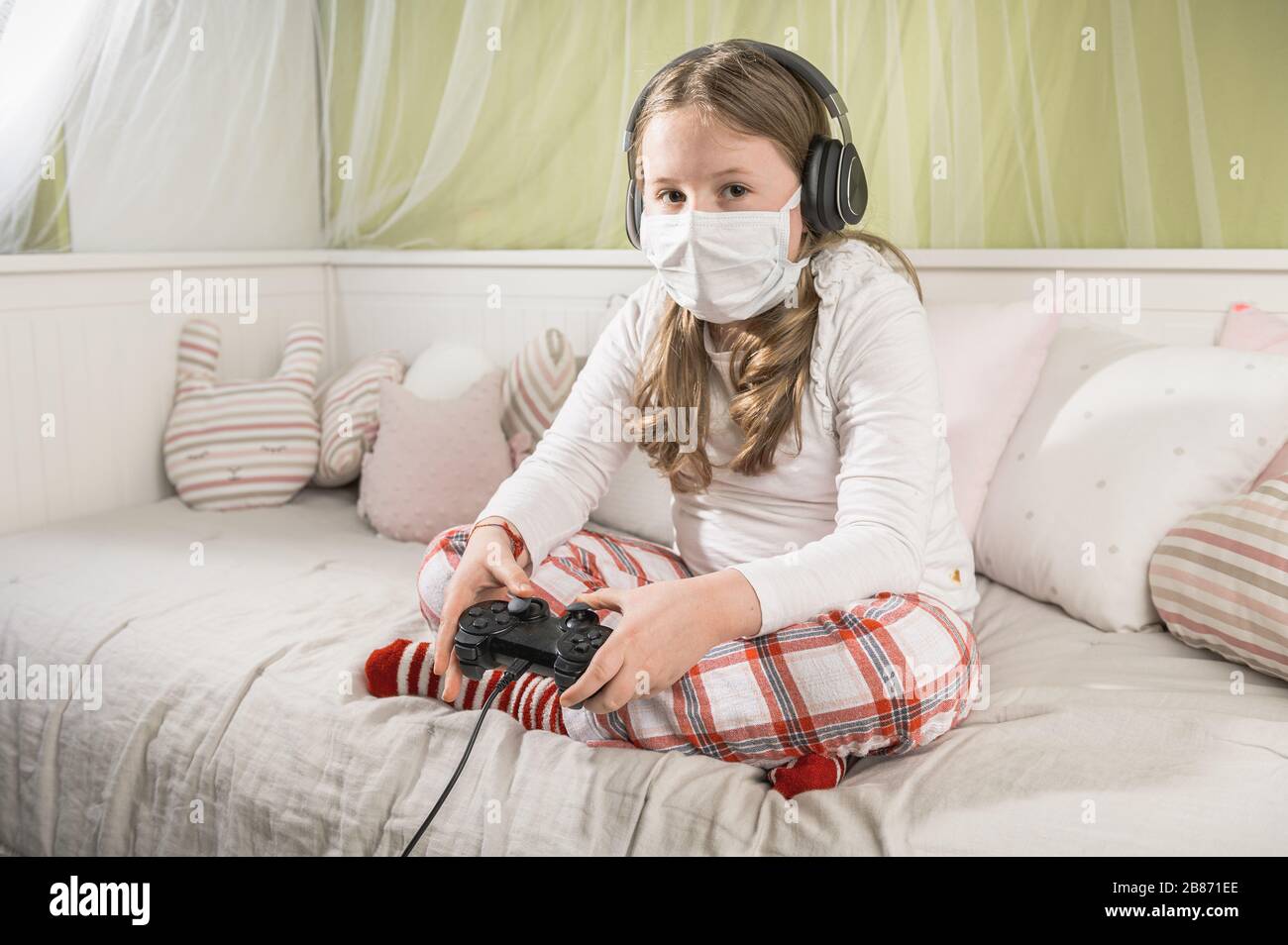 Girl in a mask playing on a game console during quarantine on the bed. Stock Photo