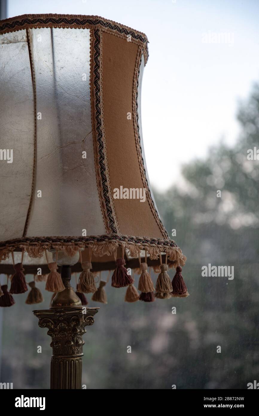 Matte transparent vintage lampshade with tassels by bottom edge in front of blurred view through window. Old-fashioned antique table lamp closeup. Stock Photo