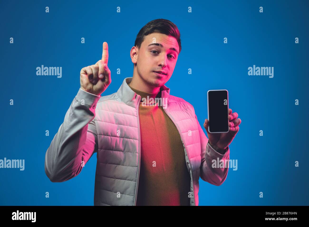 Showing blank phone screen Caucasian man's portrait on blue studio background in pink neon light. Beautiful male model in casual. Concept of human emotions, facial expression, sales, ad. Copyspace. Stock Photo