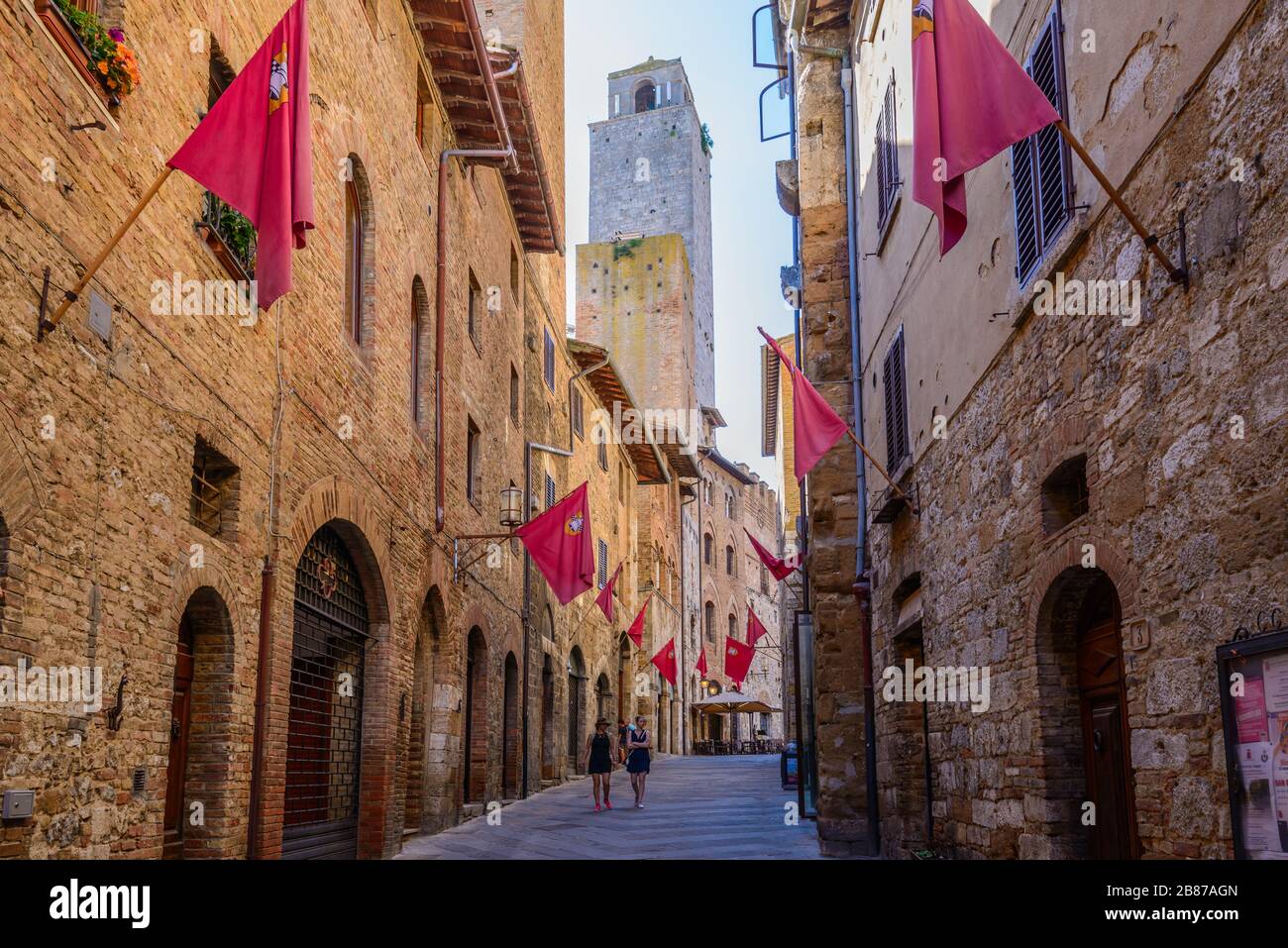 Two women walking down Via S. Matteo street with red flags flying on the walls in the historic centre of San Gimignano, Province of Siena, Italy. Stock Photo