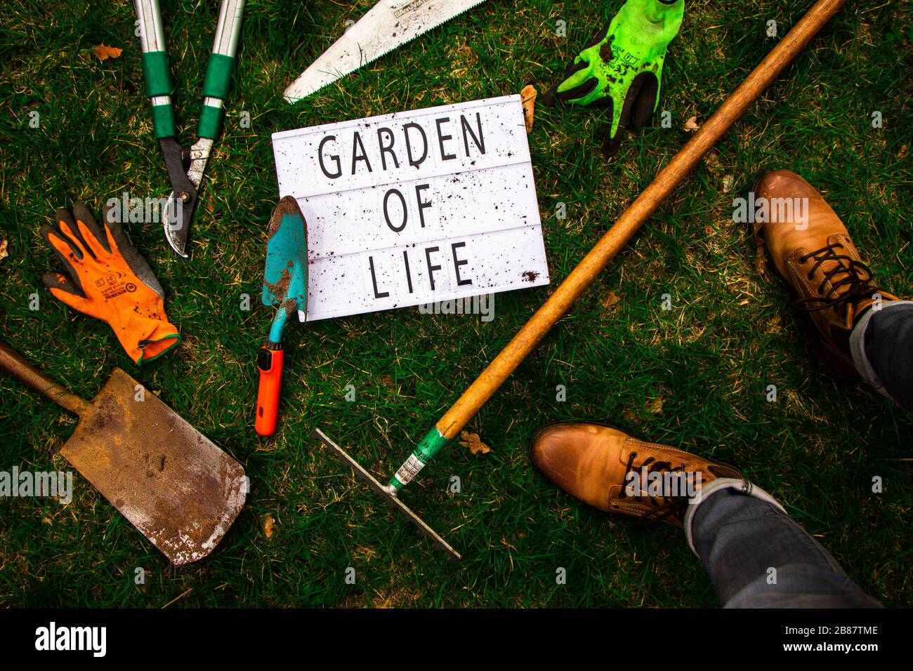 Top down view of a lightbox surrounded by tools of a gardener and the garderner himself. The lightbox has 'Garden Of Life' spelled out. Lifestyle. Stock Photo