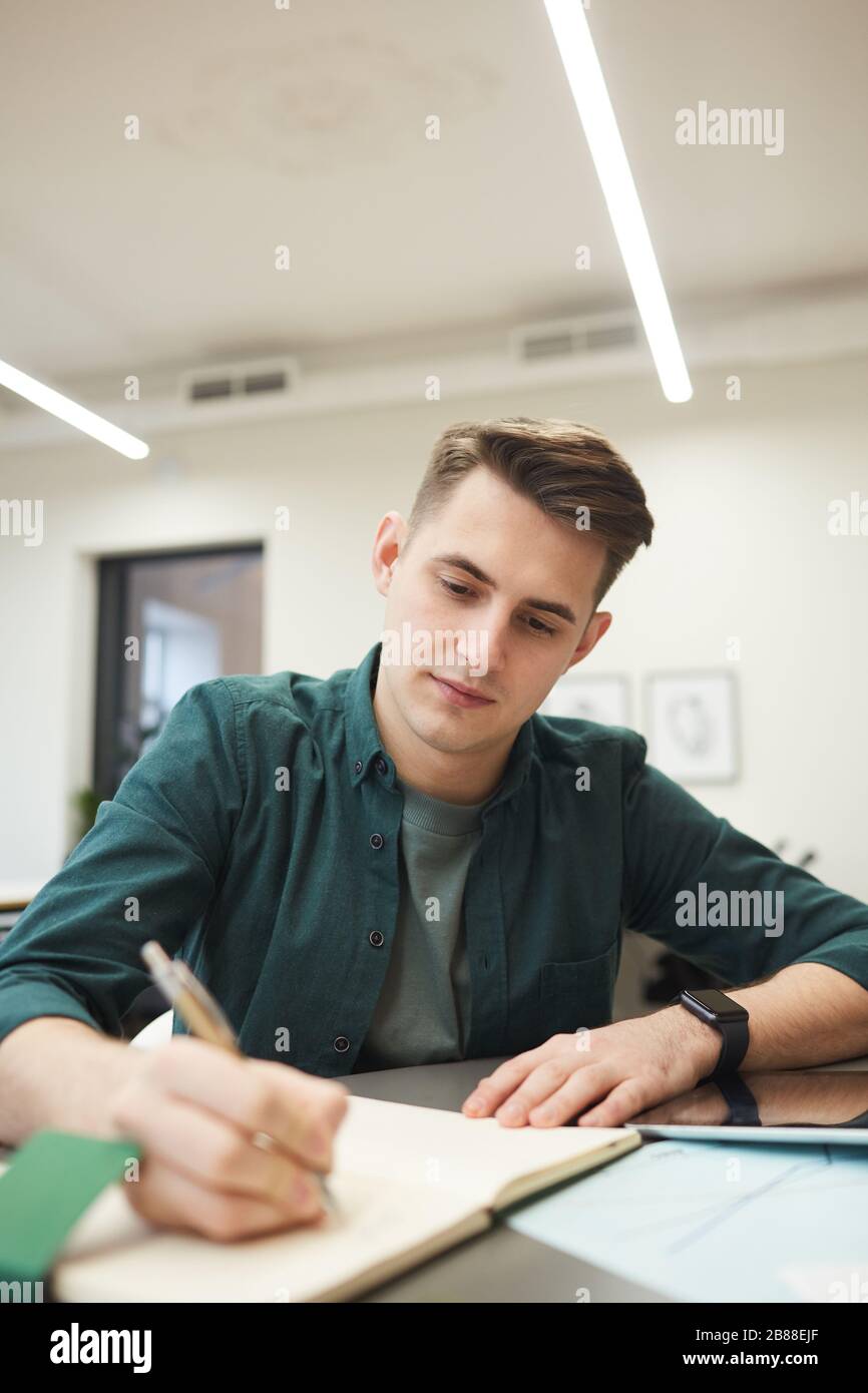 Young man sitting at the table and making notes in notepad while working at office Stock Photo