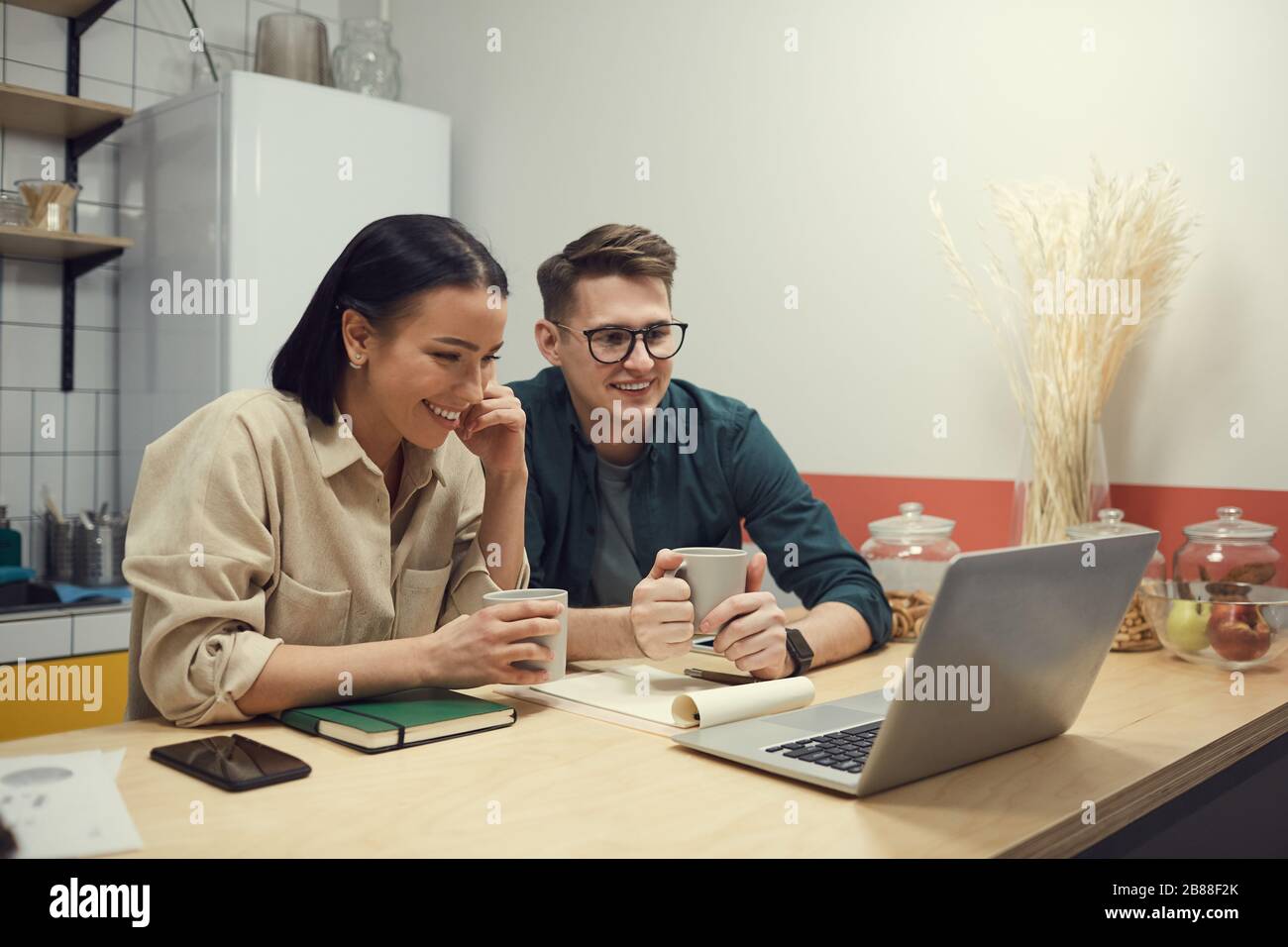 Two business partners sitting at the table drinking coffee and communicating online with their colleague on laptop computer in the kitchen Stock Photo
