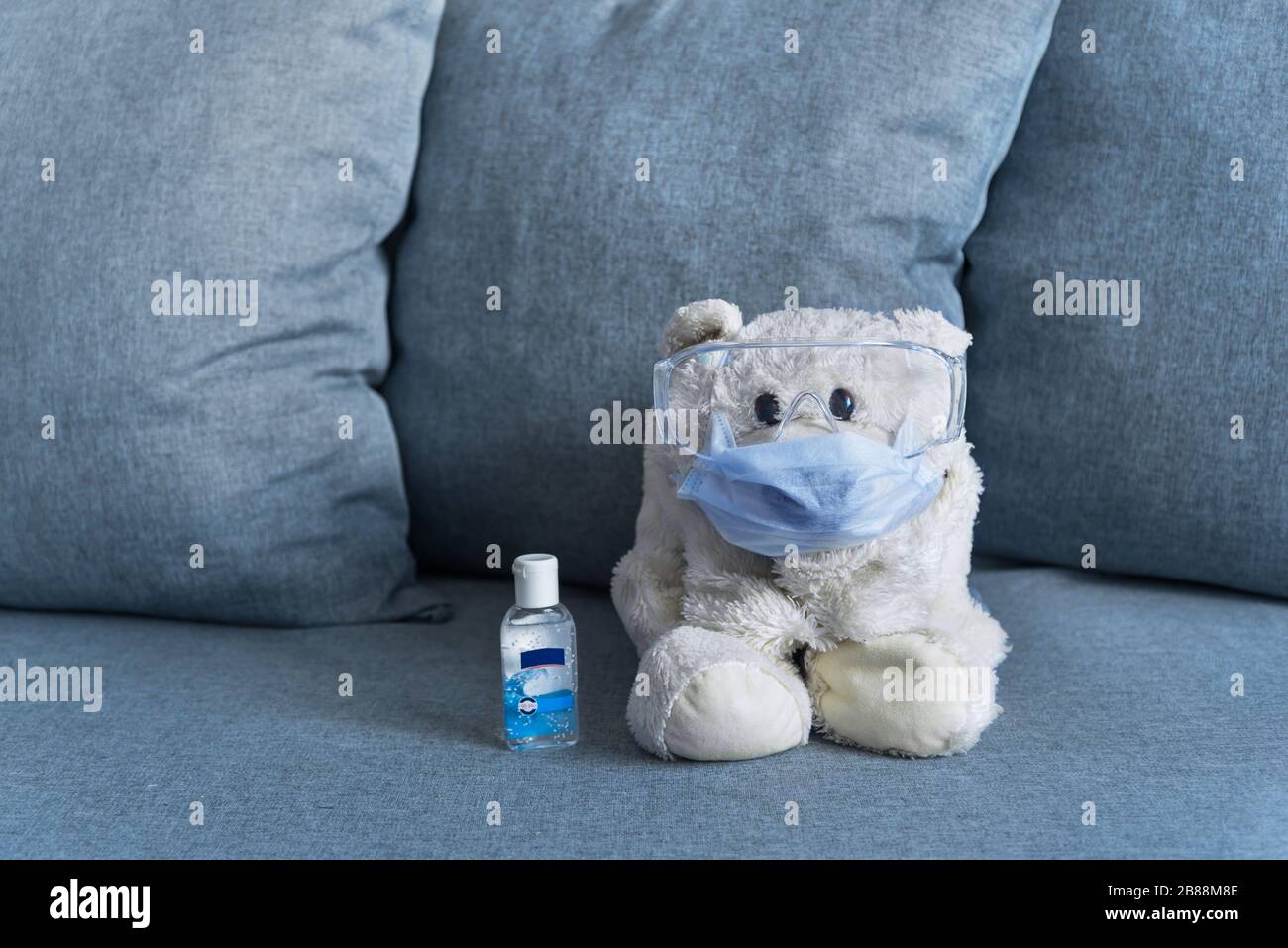 white teddy bear sitting on sofa wearing mask and protective glasses with hand sanitizer next to it Stock Photo