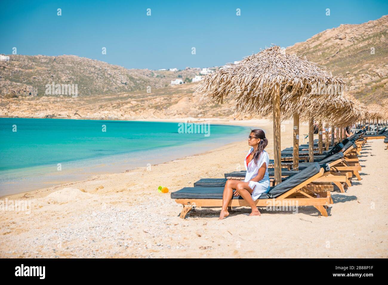 woman vacation Mykonos beach during summer with umbrella and luxury beach chairs beds, blue ocean with mountain at Elia beach Mikonos Greece Stock Photo
