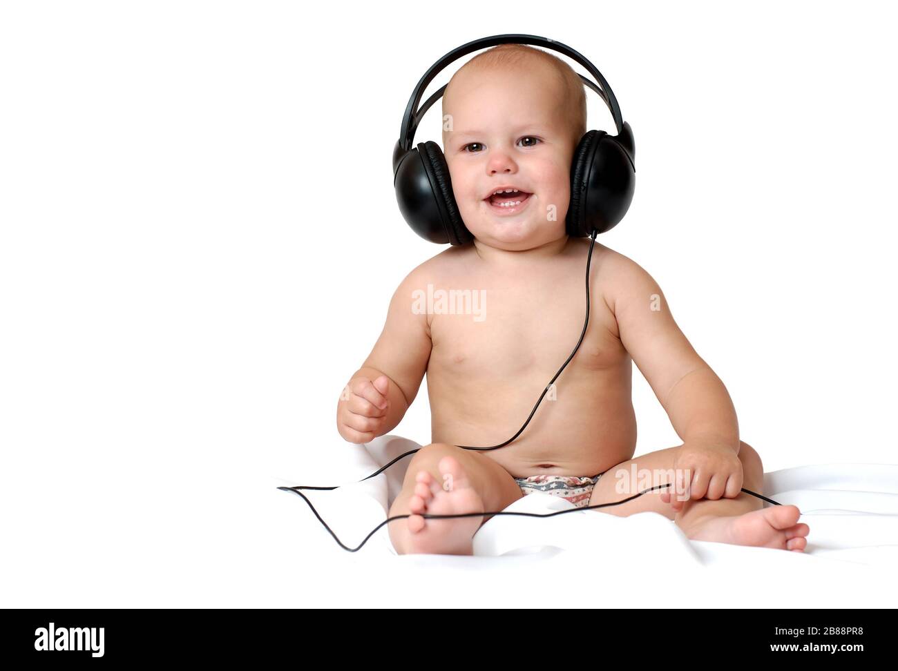 Little one year old boy listens to music in big headphones and smiles on an isolated white background Stock Photo