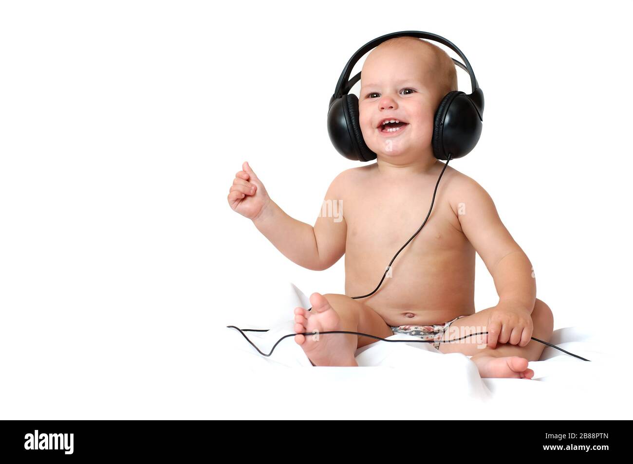 Little one year old boy listens to music in big headphones and smiles on an isolated white background Stock Photo