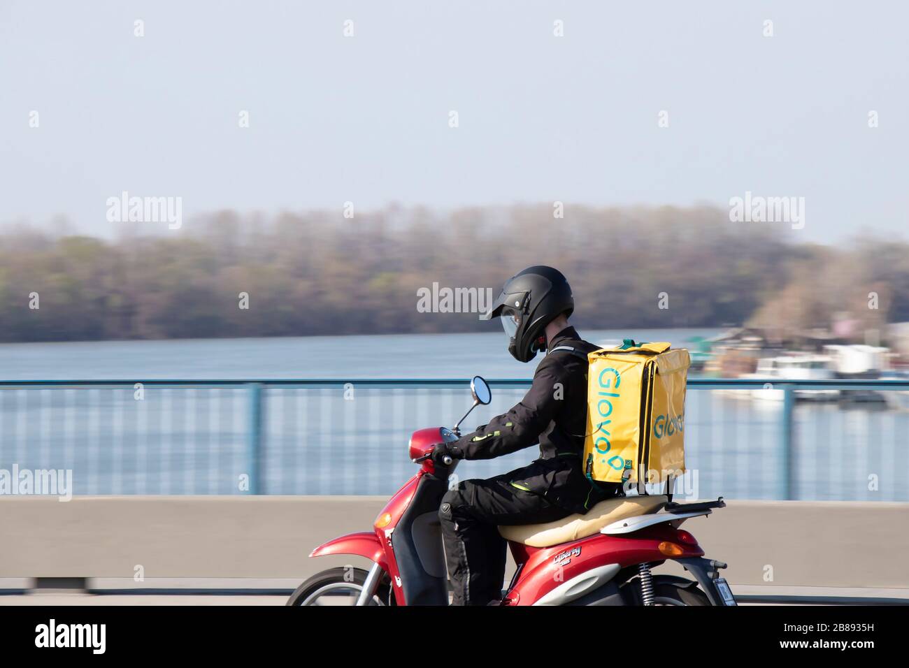 Belgrade, Serbia - March 18, 2020 : A courier working for Glovo food delivery service riding a scooter bike on the bridge over the Sava river Stock Photo
