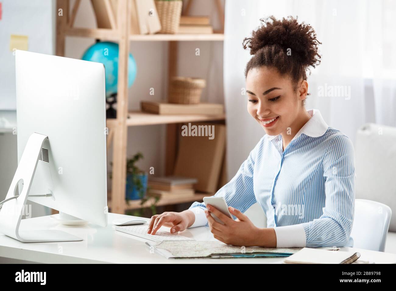 Tour agent sitting at table in office browsing computer looking at new message on smartphone happy Stock Photo