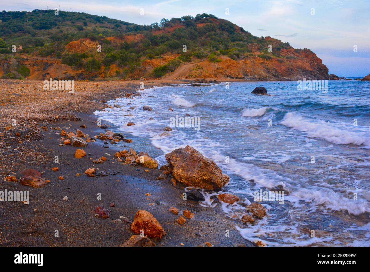 A beautiful rocky coast with color contrast: orange stones and vulcanic black sand. Strong sea waves and little hill with pine trees in the back. Stock Photo