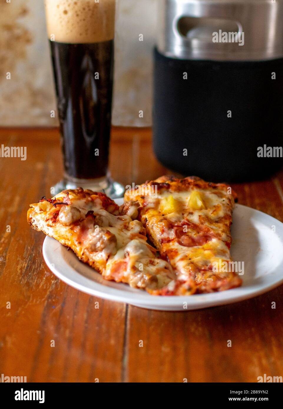 two slices of hot, gooey pizza are paired with a tall glass of beer and a mini growler Stock Photo