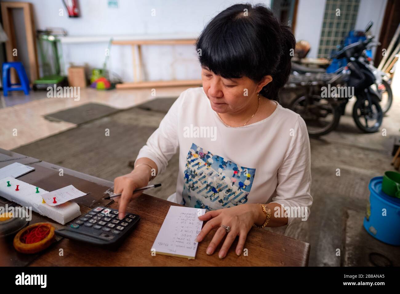 Asian Indonesian women, the owner of small local family-owned business store, or locally called warung, calculating profit by her desk. Selective Focu Stock Photo