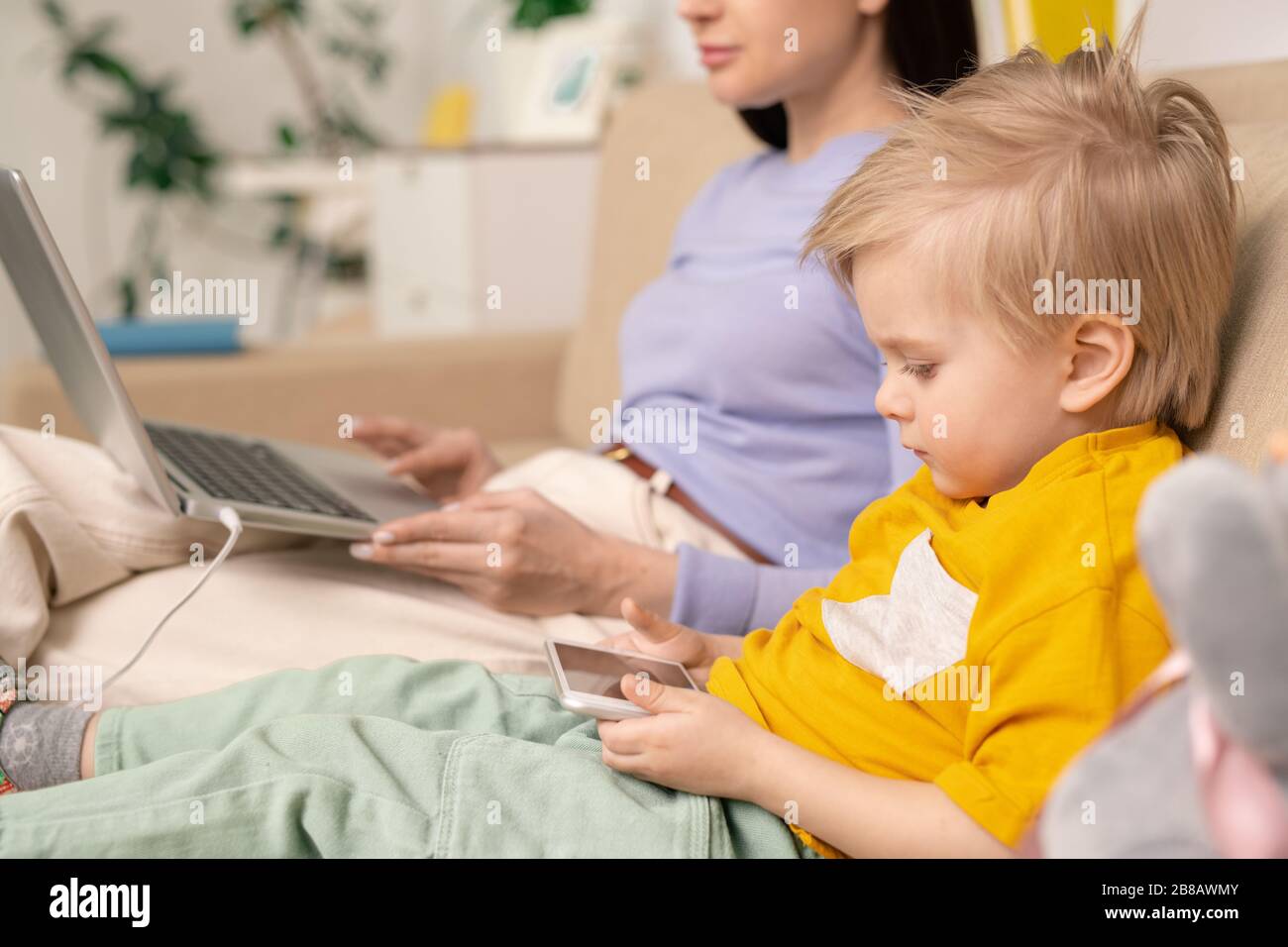 Close-up of serious concentrated kid boy sitting on sofa and using gadget while mother working with laptop Stock Photo