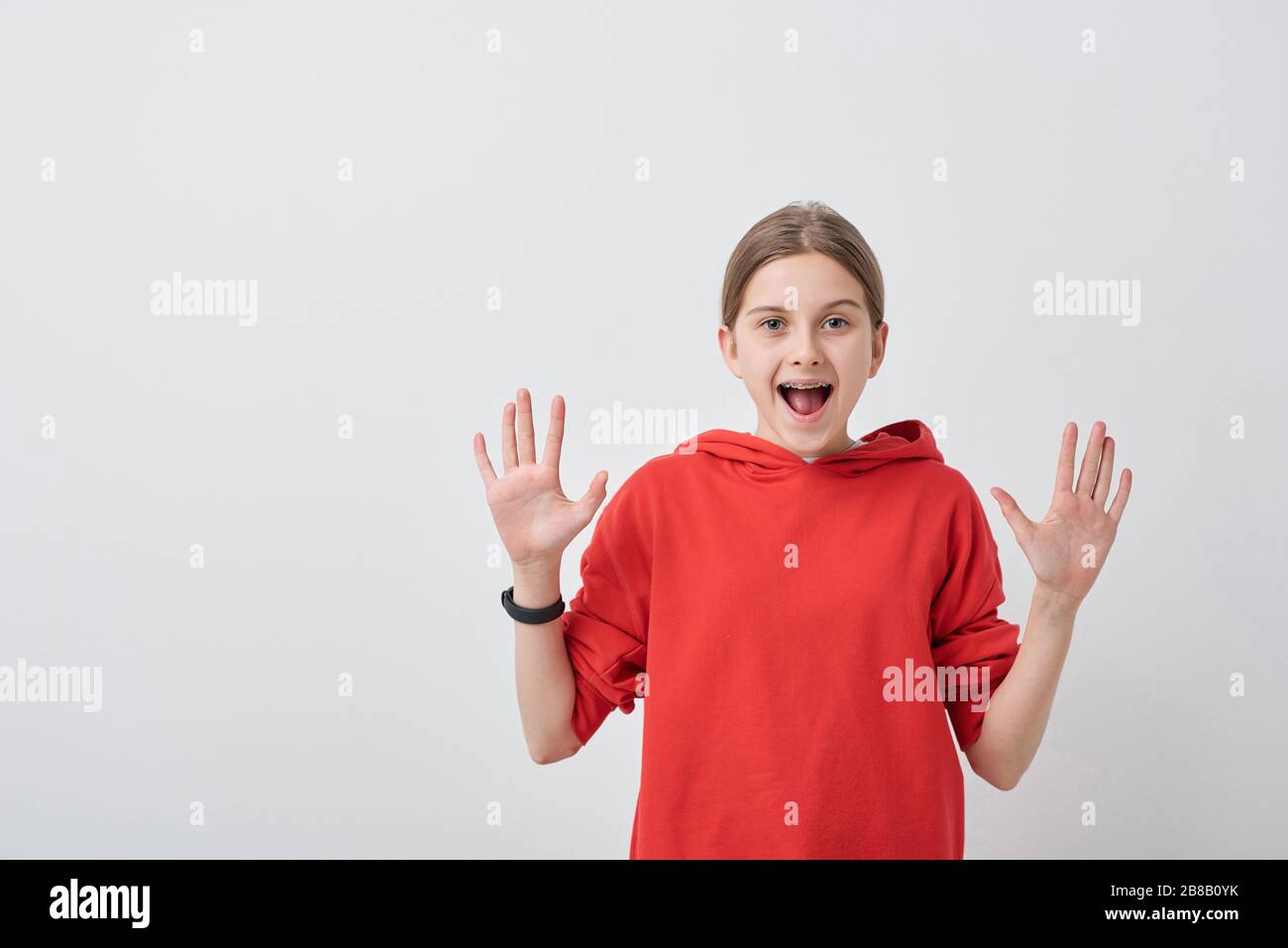 Friendly and cheerful teenage girl in red sweatshirt looking at you with her palms open and expressing gladness over white background Stock Photo