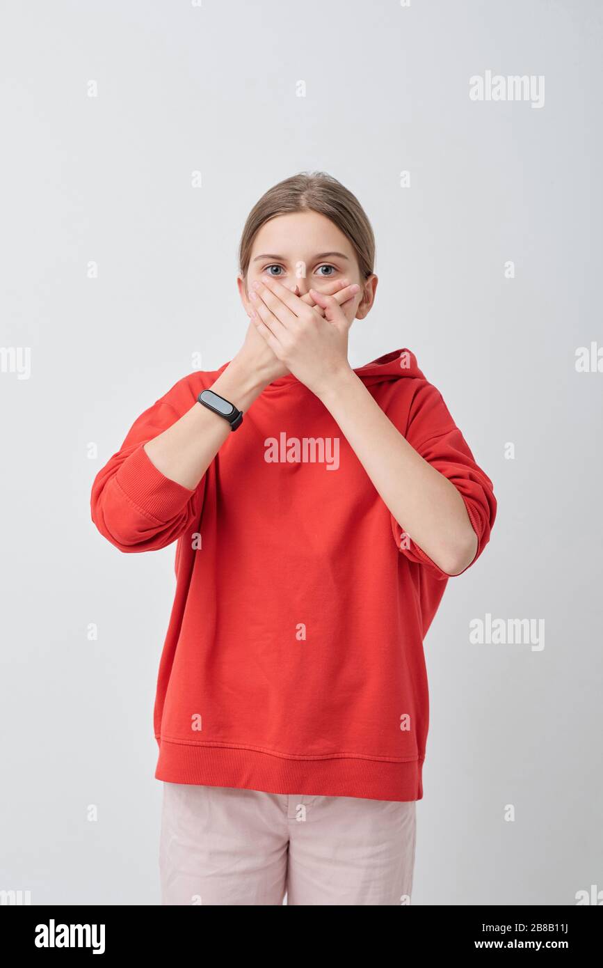 Pretty teenager in red hoodie and white jeans covering her mouth by hands while expressing reluctance to tell a secret in isolation Stock Photo